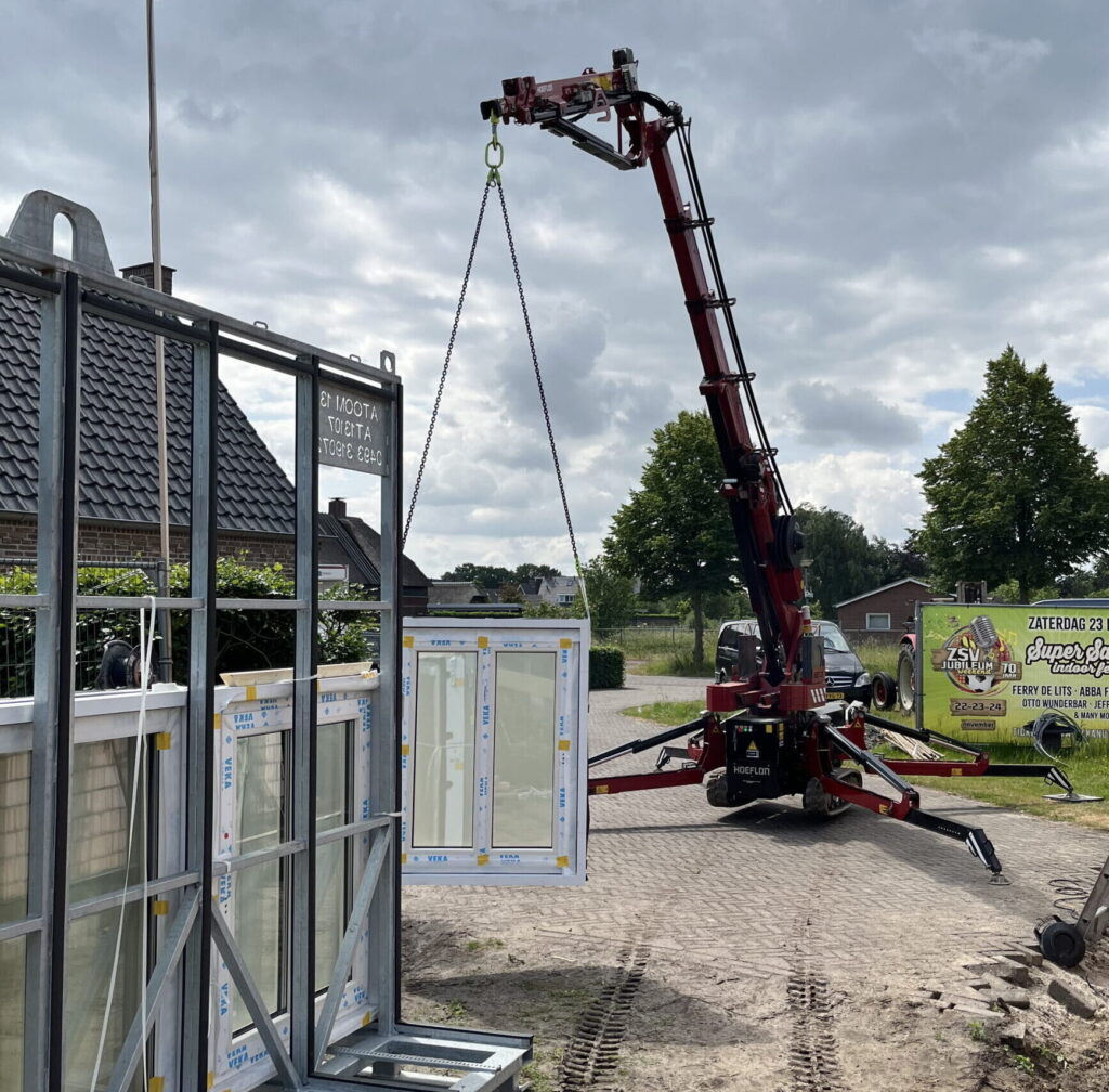 Een kleine hijskraan die een prefab kozijn van een stalen bok af hijst.